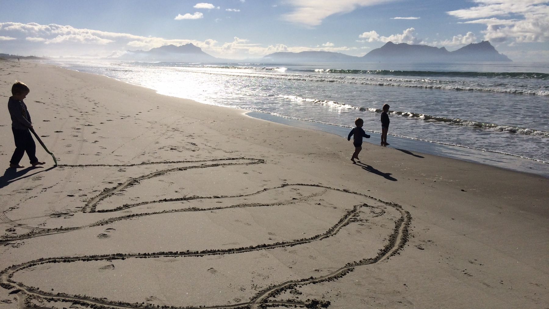 Three children on the wet sand at Bream Bay early in the morning, with a large four-blade propeller drawn in the sand beside them and the Whangarei Heads headlands across the water